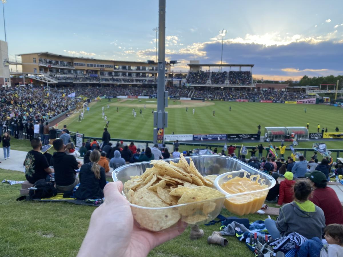A white hand holds a container of tortilla chips and cheese. In the background is the New Mexico United pitch, with players playing soccer.