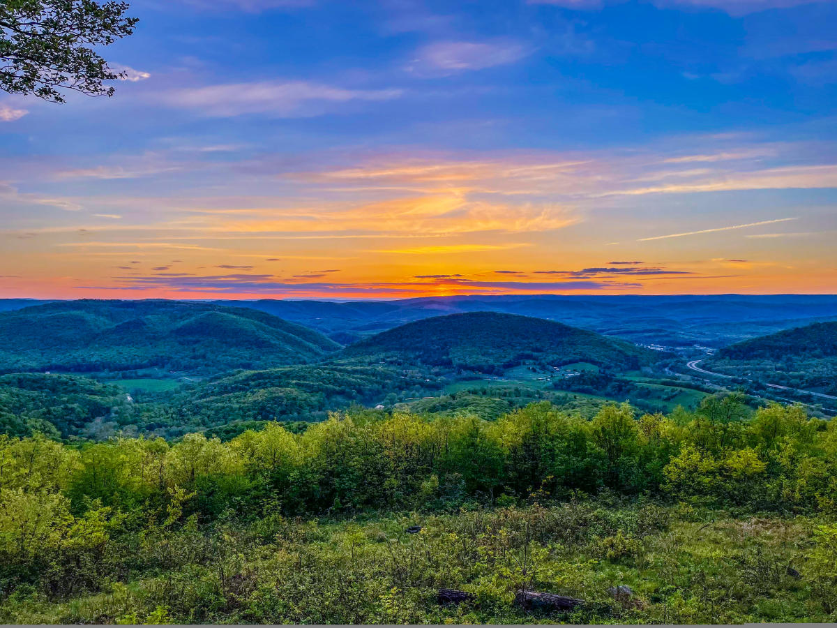 The sky is painted in orange, yellow, purple and blues as the sun sets over the mountains along the grassy mountain edge.