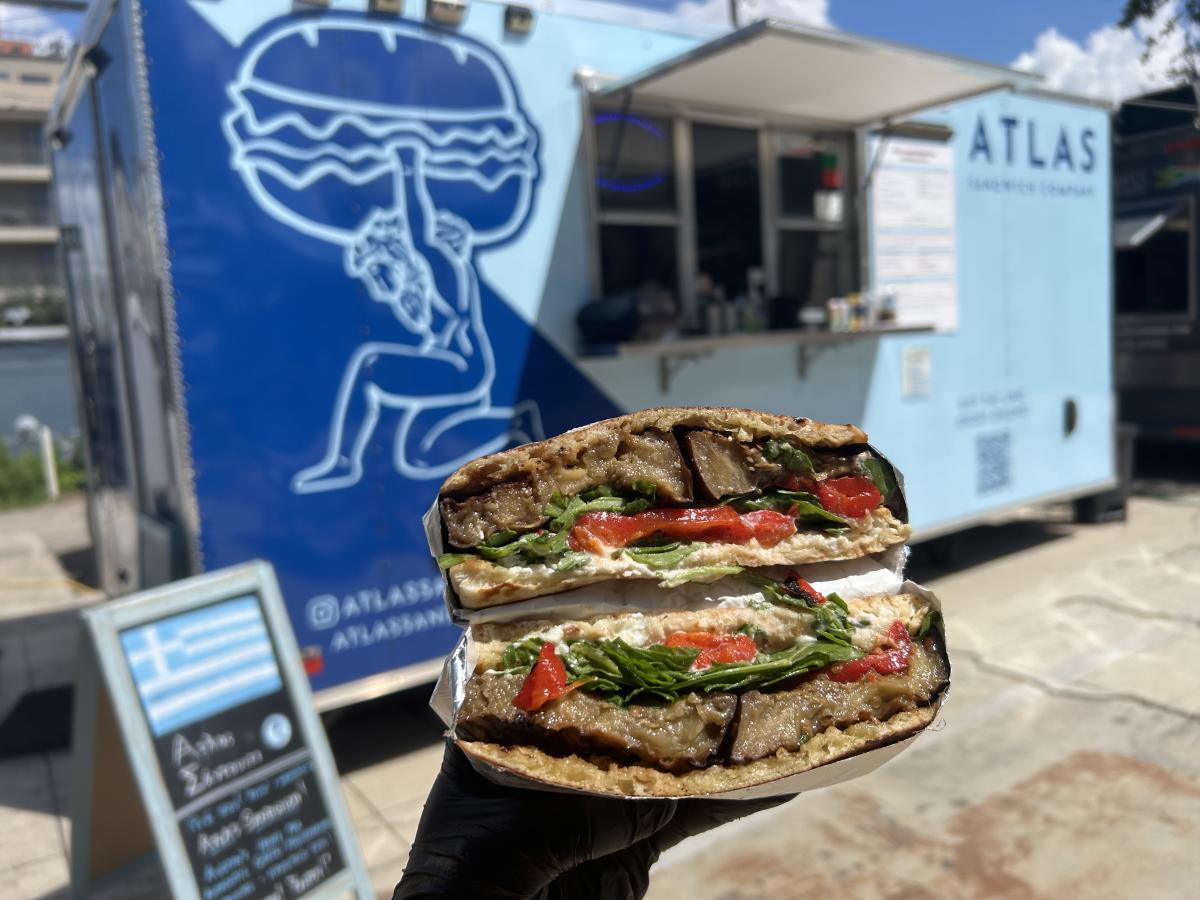 Image of a hand holding a sandwhich that's been sliced in half with the Atlas Sandwich Co. food truck in the background.