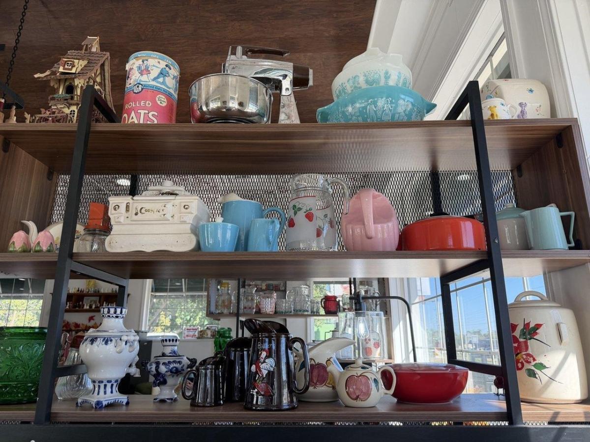 Wooden shelves with black metal supports display vintage kitchenware, including turquoise and pink pitchers, patterned teapots, blue-and-white ceramics, a red casserole dish, a metal mixer, and an old rolled oats tin. Sunlight streams through tall windows beside the colorful collection.