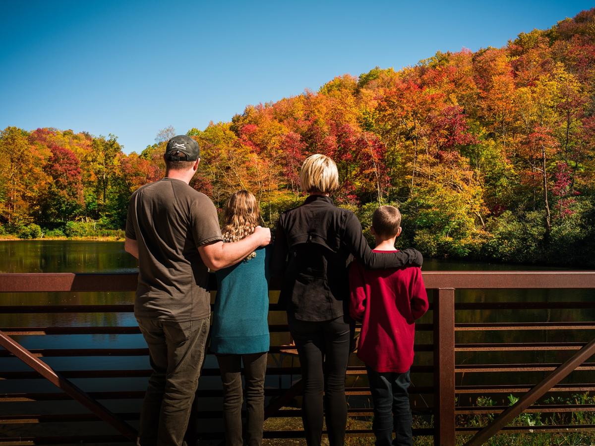A family looks over a pond towards a vibrant autumn mountainside from a rust-colored bridge.