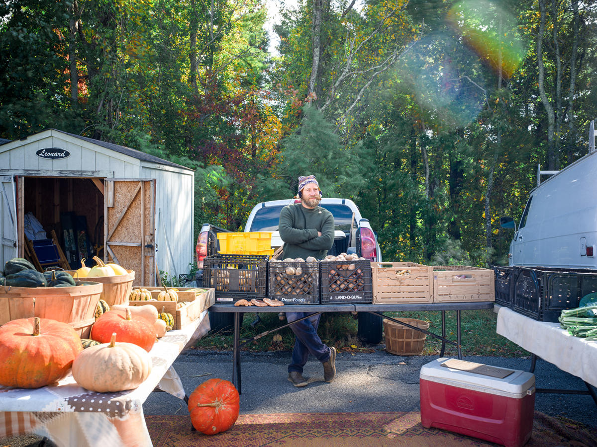 A vendor stands behind a table full of fall vegetables like pumpkins and potatoes.
