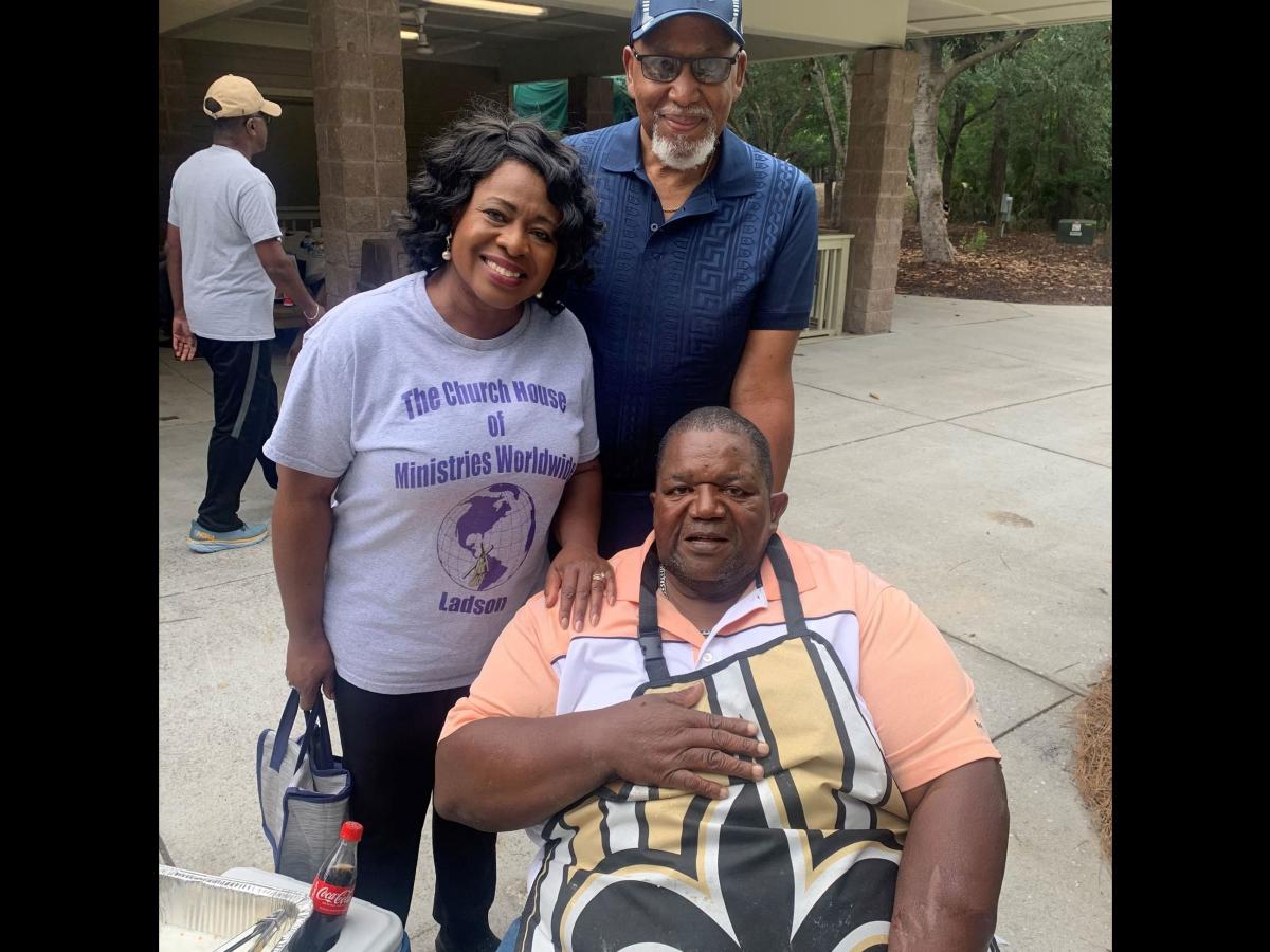 Three people pose for a picture outside near a covered shelter.