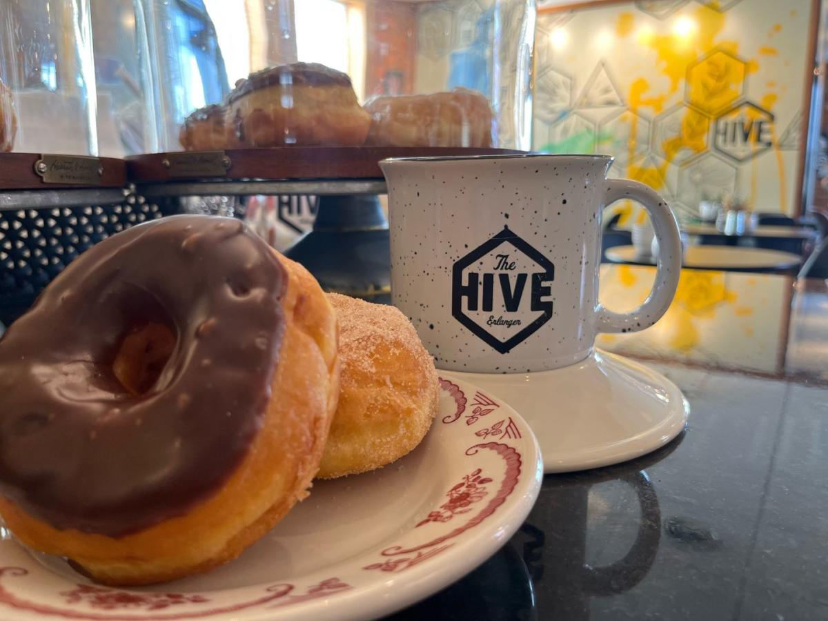 Image is of a chocolate glaze donut on a plate with a coffee mug that says "The Hive" on it.