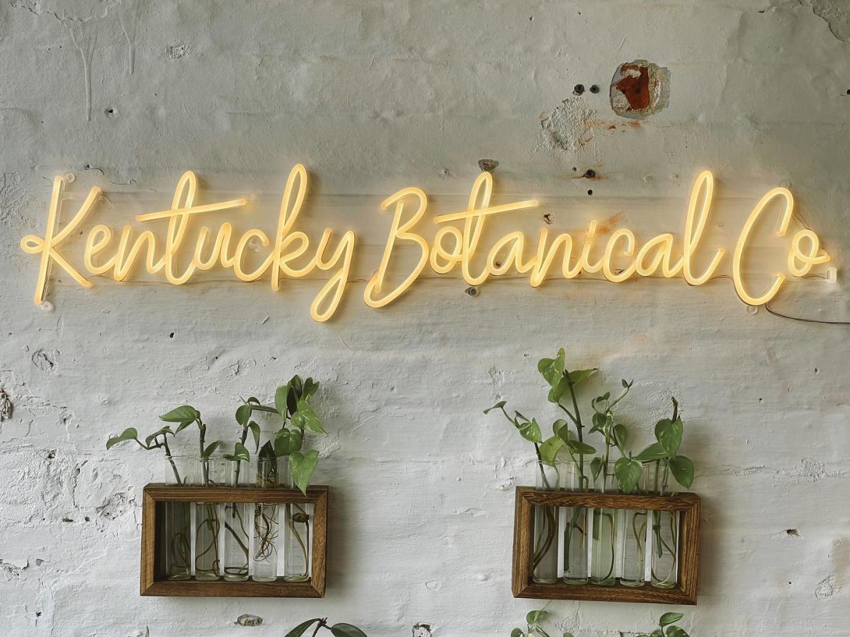 Image is of a white, painted brick wall with a neon sign that say's Kentucky Botanical Co.