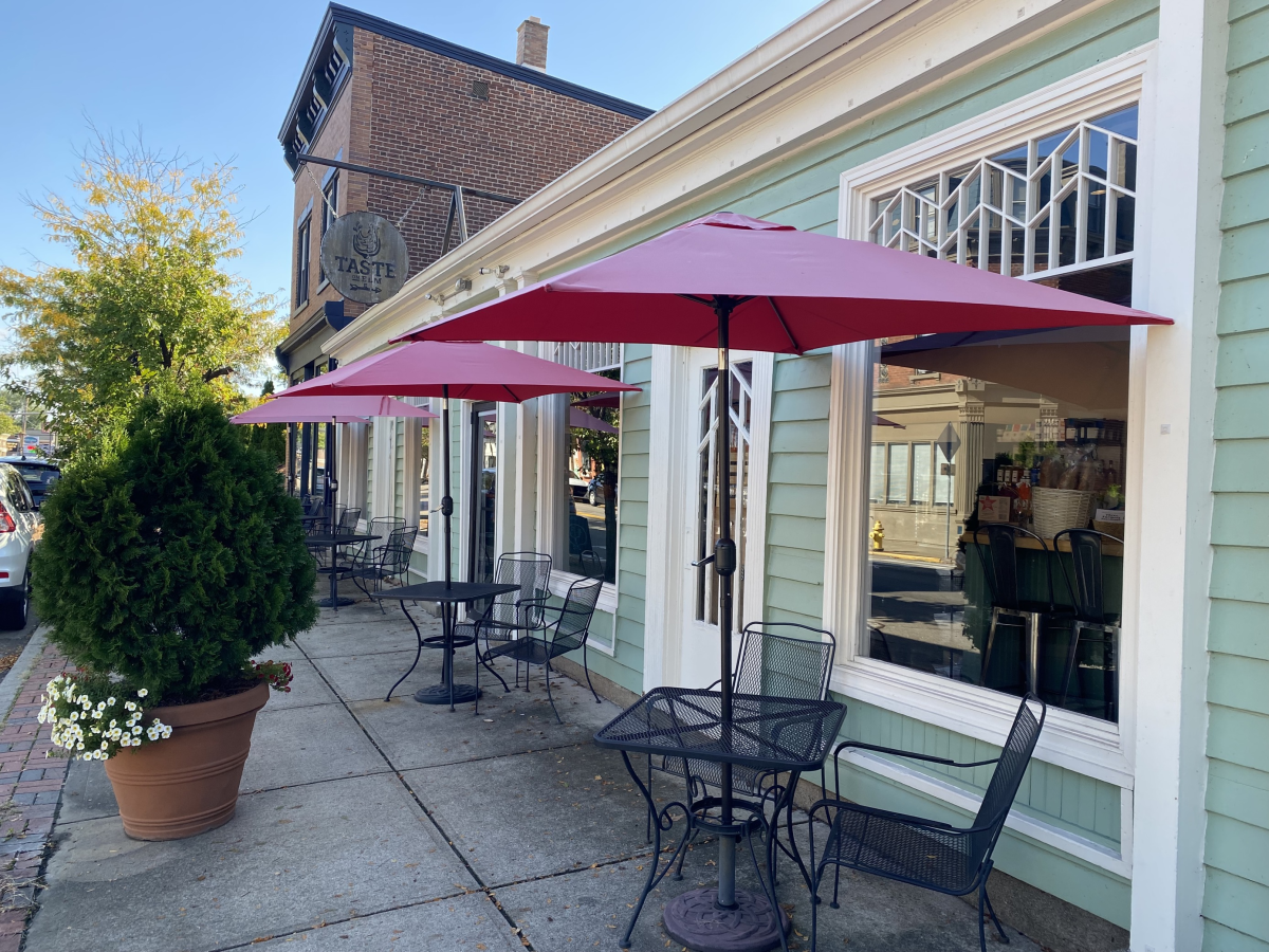 Image is of the outside facade of Taste of Elm with tables, chairs and umbrellas in front on the side walk.