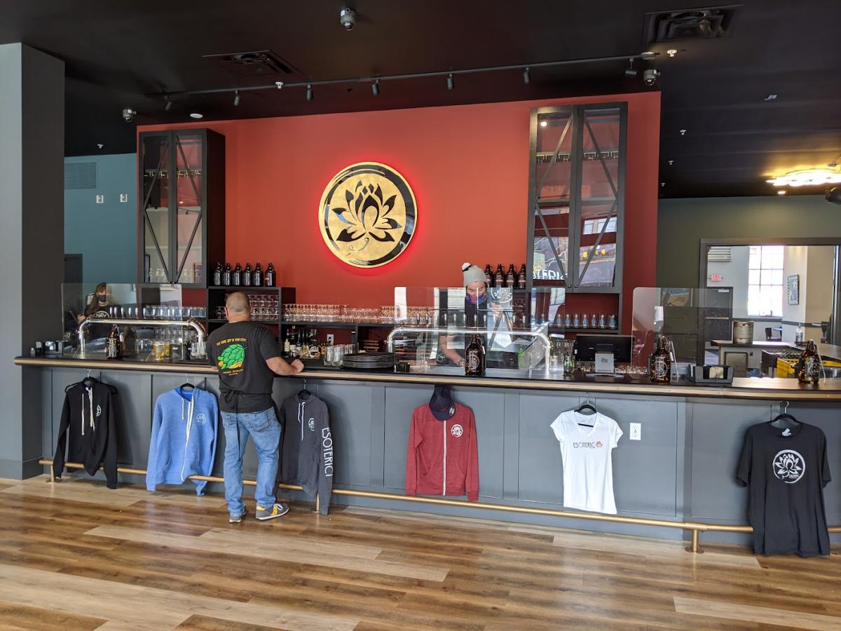 A man waiting for his order at the bar of Esoteric Brewing's taproom with a red wall bearing the Esoteric logo in the background