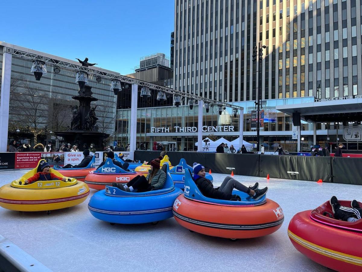 5 ice rink bumper cars colored red, blue, yellow, and orange, holding people who are laughing and bumping the cars into each other on Fountain Square in Cincinnati