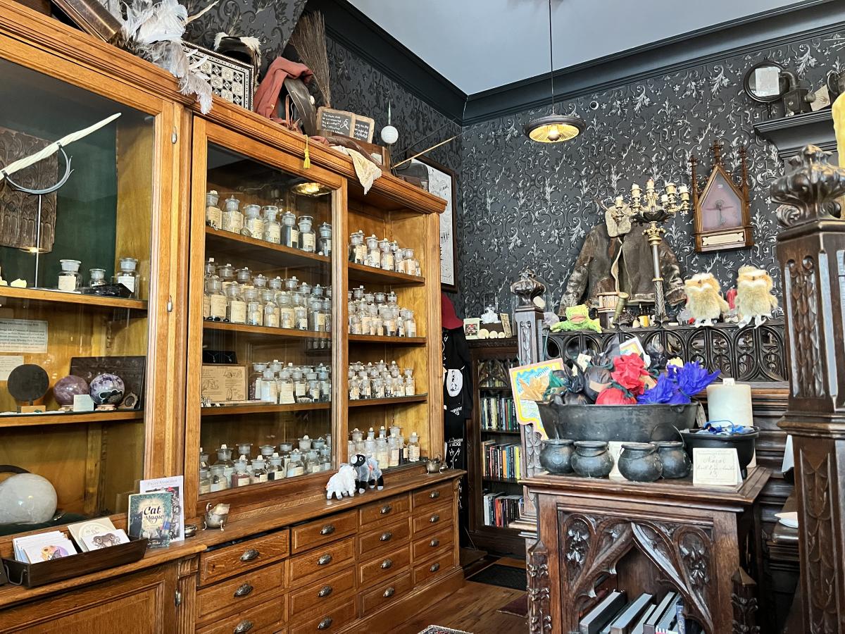 Image is of inside the store with old time bookshelves and drawers on the left with vials and stones and on the right is more freestanding shelves with a caldron and wands.