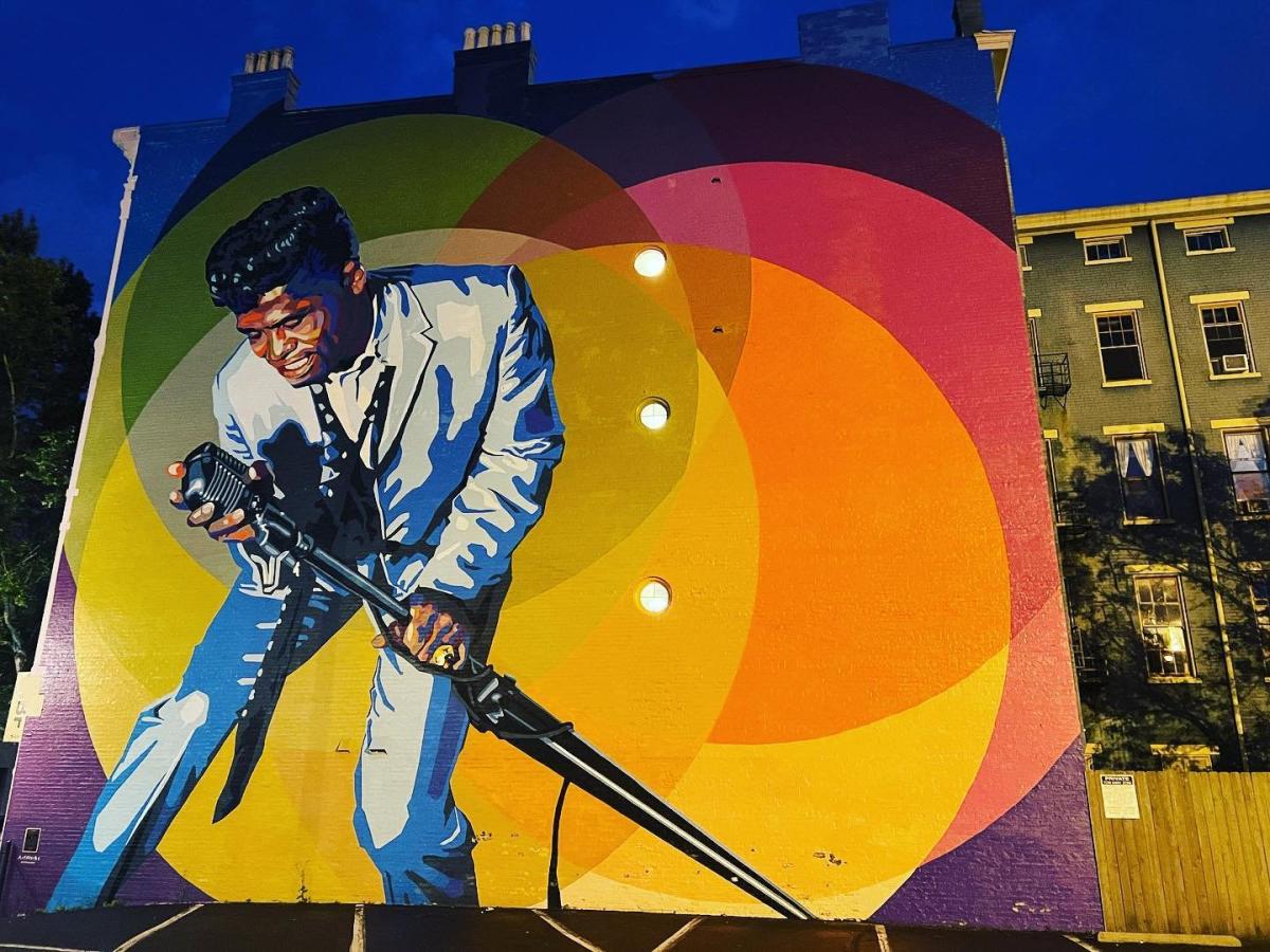 A mural of James Brown holding a microphone and leaning over to sing with yellow orange and red circles in the background