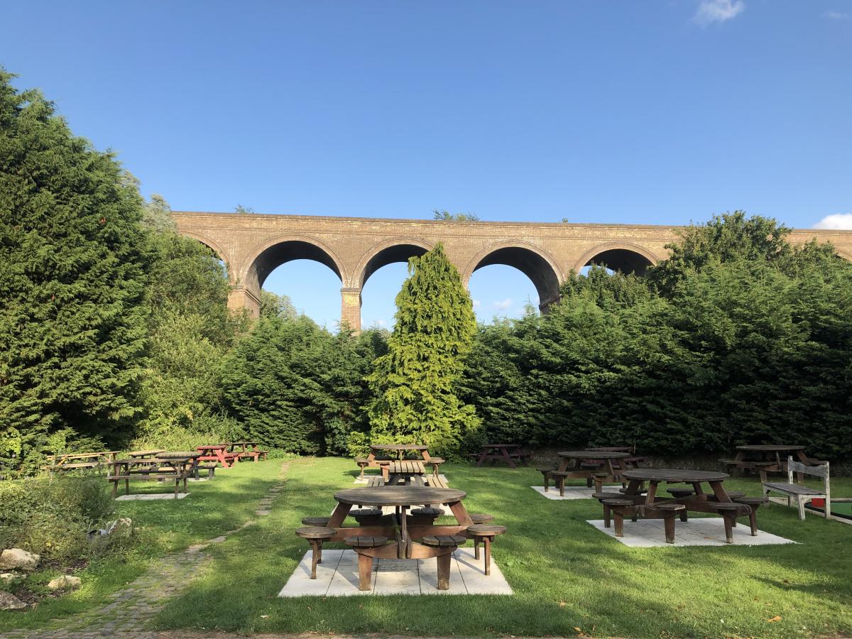 Wooden Tables sit on a lawn in a pub beer garden surrounded by greenery. A large, picturesque brick viaduct sits behind them in the middle distance