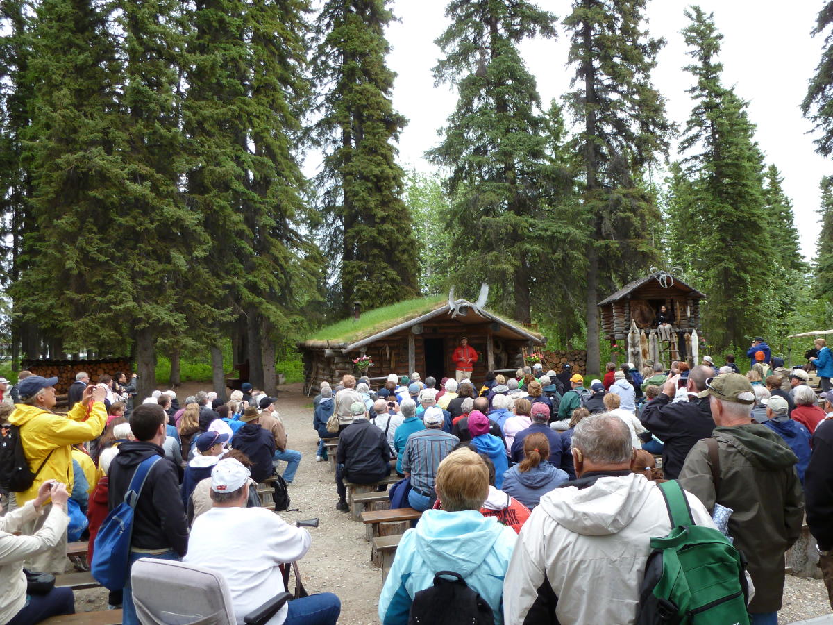 A large group of people gather and listen to a presentation on Alaska Native Culture in the Chena Village along the Chena River.
