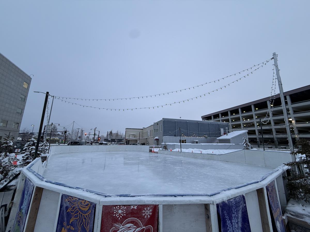 A shiny ice rink in downtown Fairbanks, AK surrounded by holiday lights, buildings, and trees.