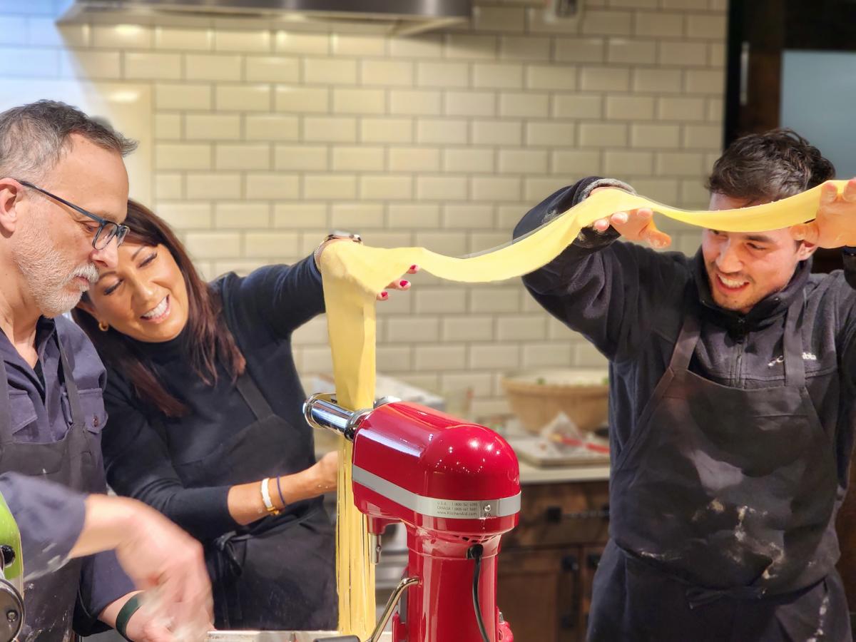 Three people using a kitchen aid to cut noodle dough into noodles