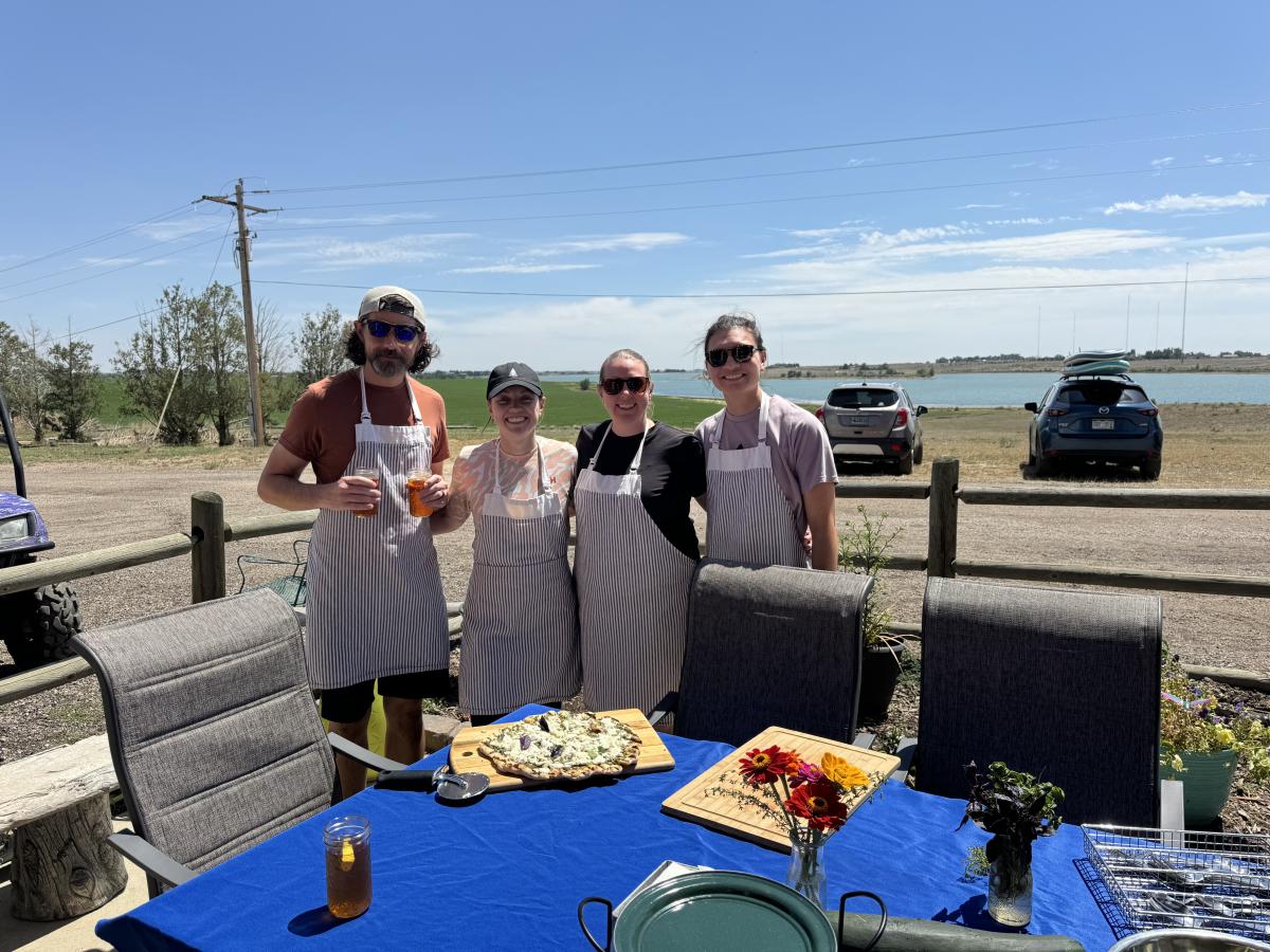 Four people standing on a patio with a lake in the background with food on the table
