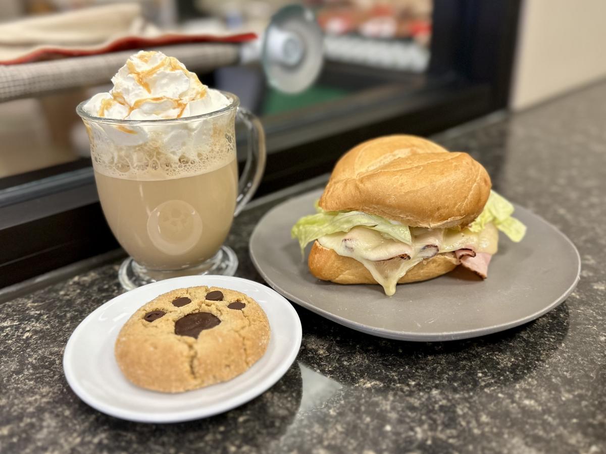 A meal consisting of a toasted sandwich with melted cheese, ham, and lettuce on a roll, served on a gray plate. Accompanying the sandwich is a peanut butter cookie with chocolate chips arranged in a paw print design on a small white plate, and a glass mug of caramel-topped latte with whipped cream.