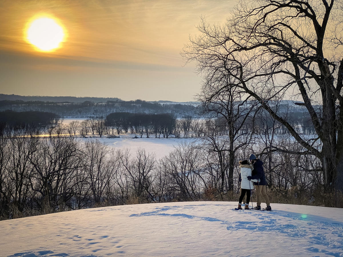 Two people in winter, looking over a bluff on a snowy hill. The sun is setting in the distance.