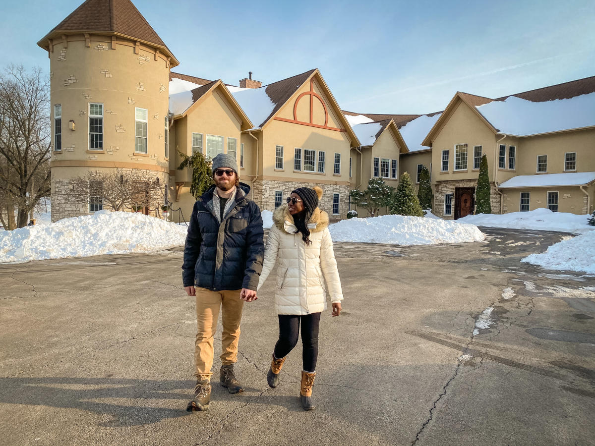 A couple holding hands, walking in the wintertime with Goldmoor Inn behind them. There is a snow in the background around the building.