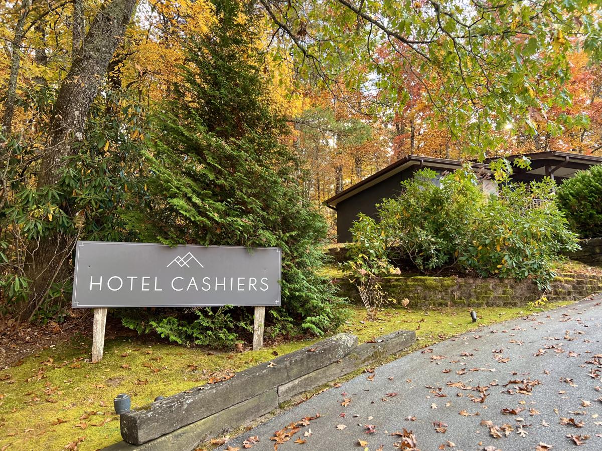 A gray sign reading "HOTEL CASHIERS" with a minimalist mountain logo stands on a mossy green lawn next to an asphalt driveway scattered with fallen brown leaves. The sign is nestled among evergreen bushes and tall trees displaying bright yellow and orange fall colors, with a dark building partially visible in the background.