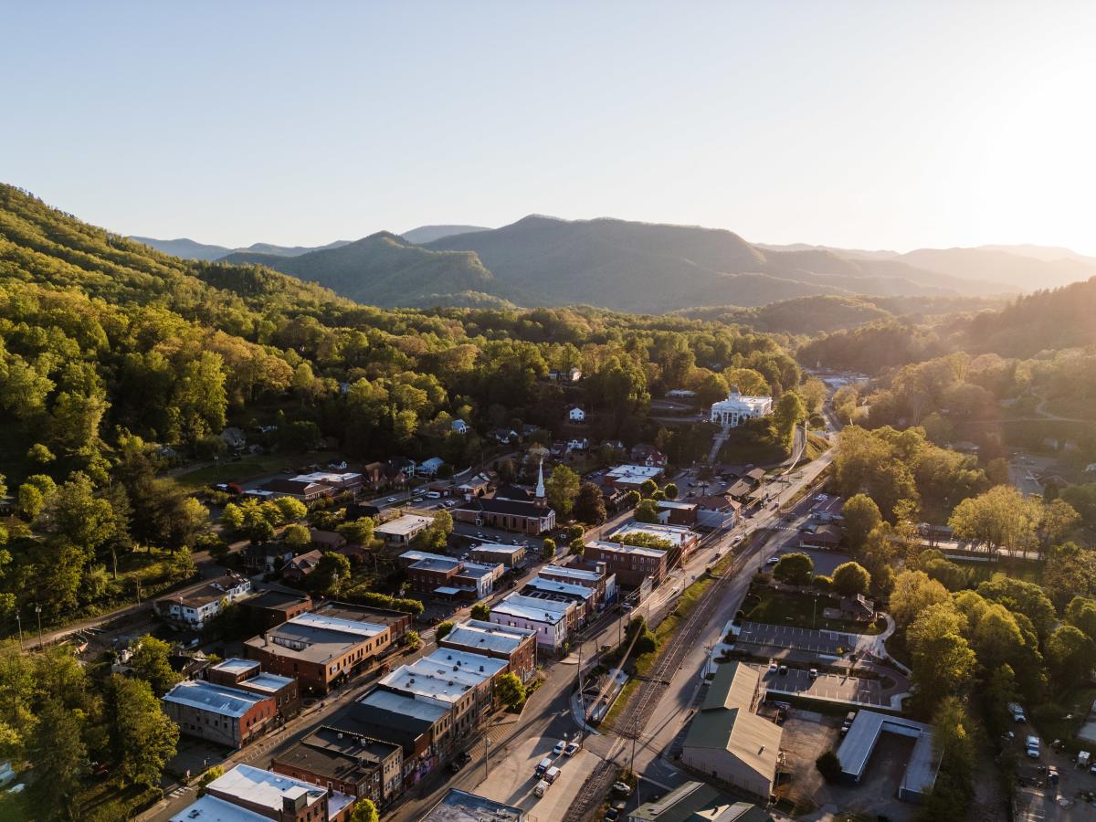 An aerial, golden-hour view of a charming small town nestled in a valley. A historic main street runs through the center, leading toward a prominent white domed courthouse situated on a hill against a backdrop of layered, forested mountains.