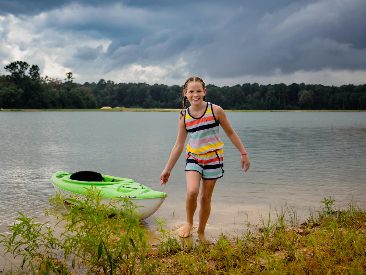 A little girl pulls a kayak out of the lake while smiling at the camera