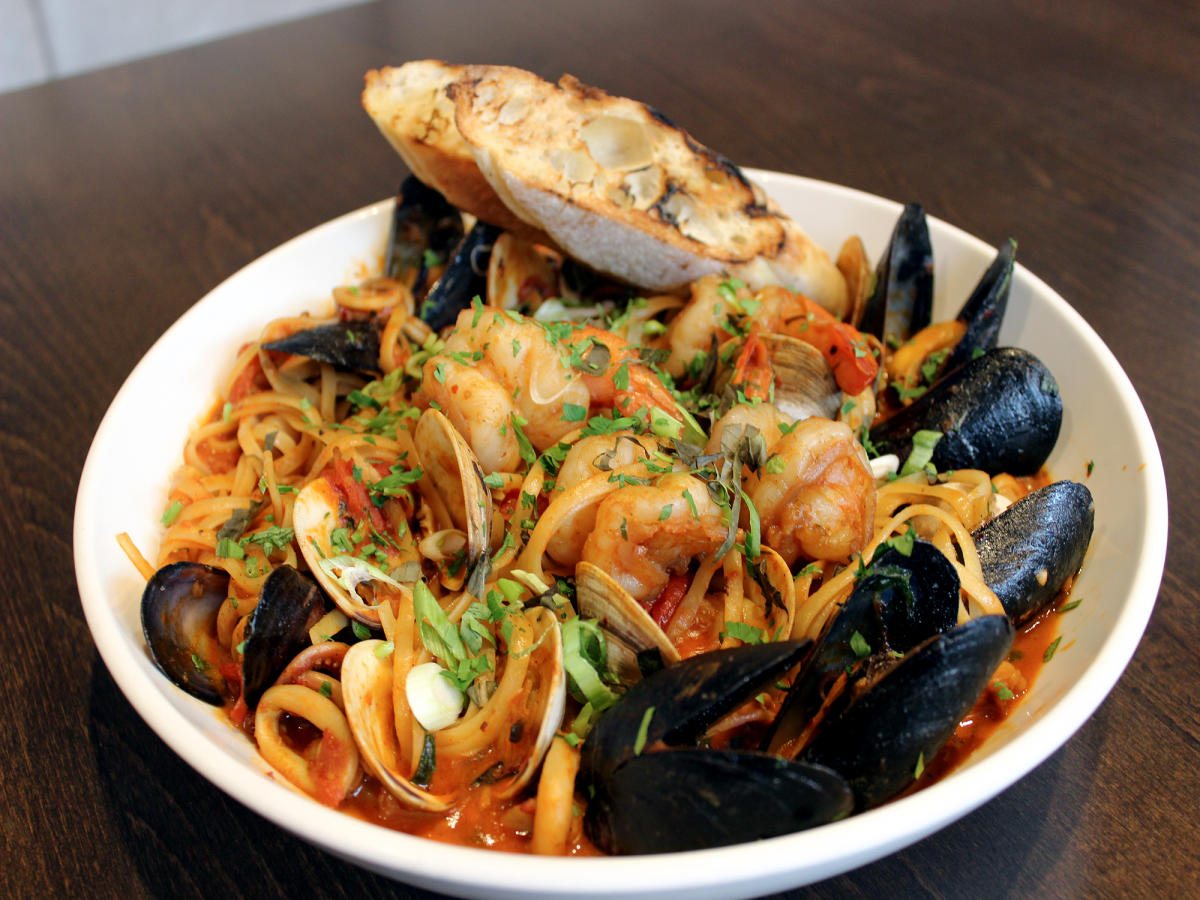 A dish with clams, mussels, shrimp, calamari, smoky tomatobroth over linguini with a side of bread.