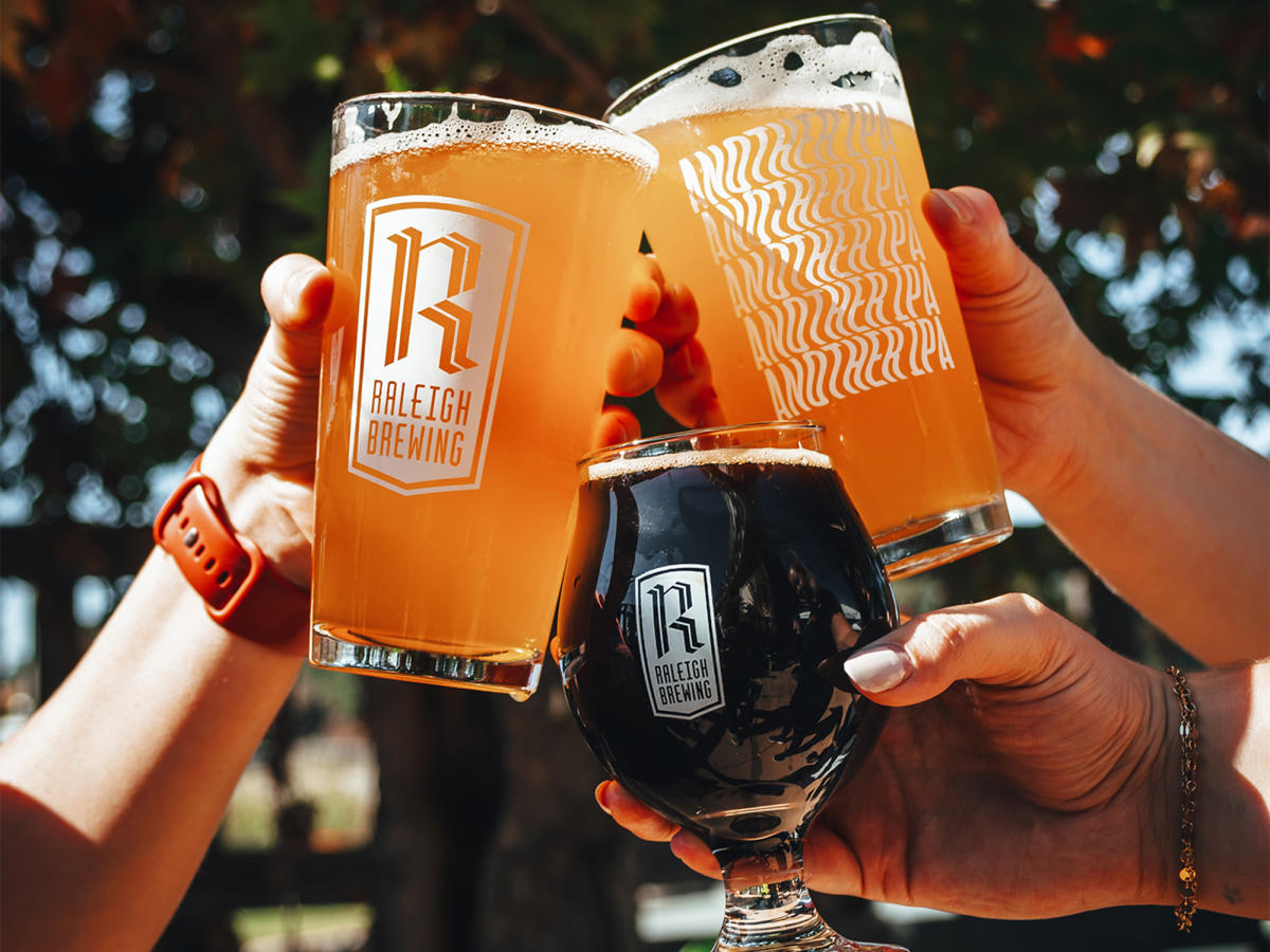 Three beers being cheers'ed by hands in the beer garden of Raleigh Brewing Distro in Smithfield, NC.