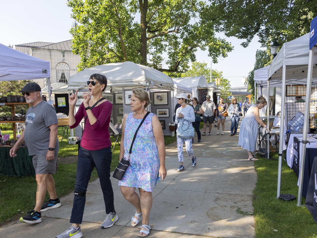 Two women and a man walk down a busy sidewalk in Bronson Park. The sidewalk is surrounded by tents with Local artisans displaying their crafted pieces.