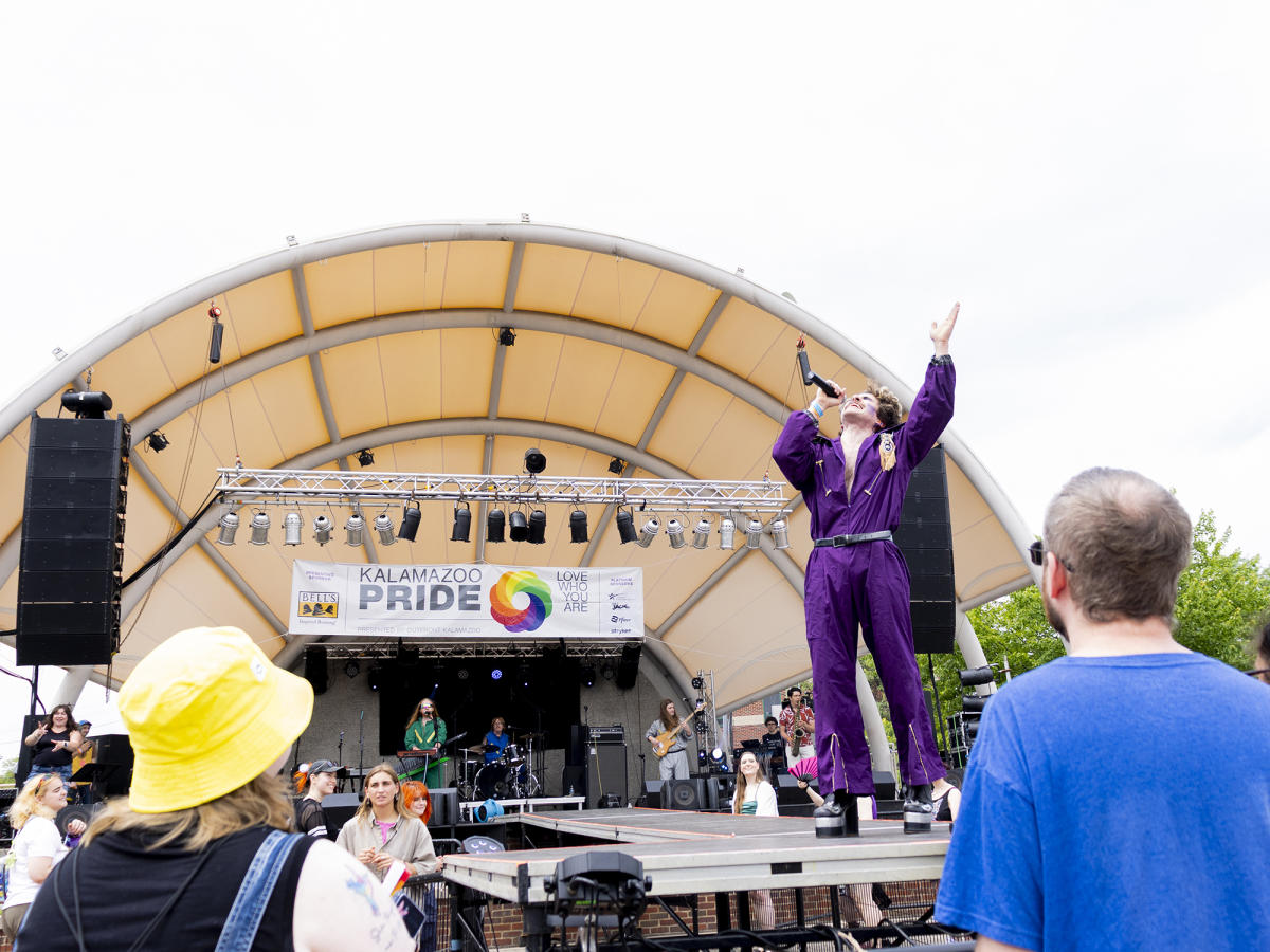 A man in a purple jumpsuit and black-heeled boots performs on an elevated stage at PRIDE in Kalamazoo, MI. A surrounding crowd enjoys and dances to his performance.
