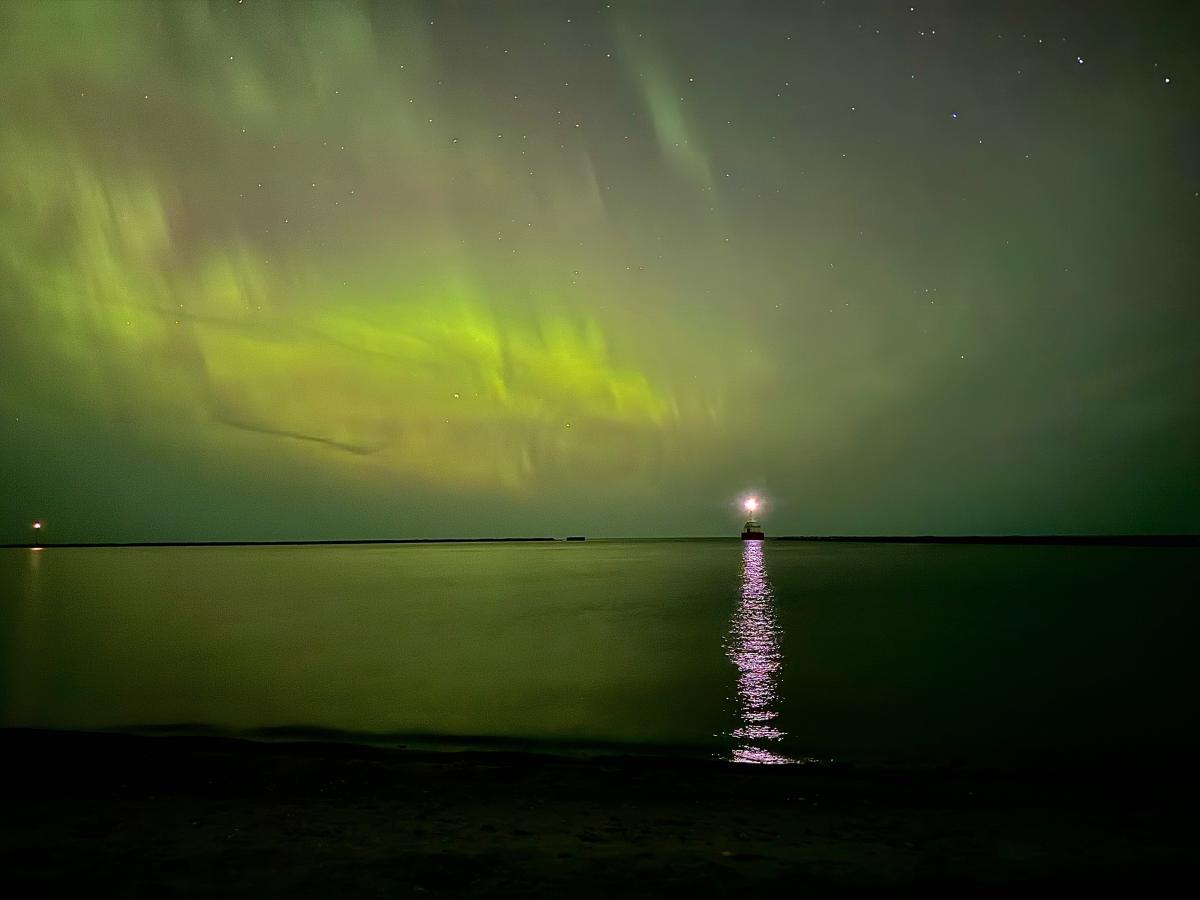 northern lights reflect off a lake, a lighthouse lit in the distance