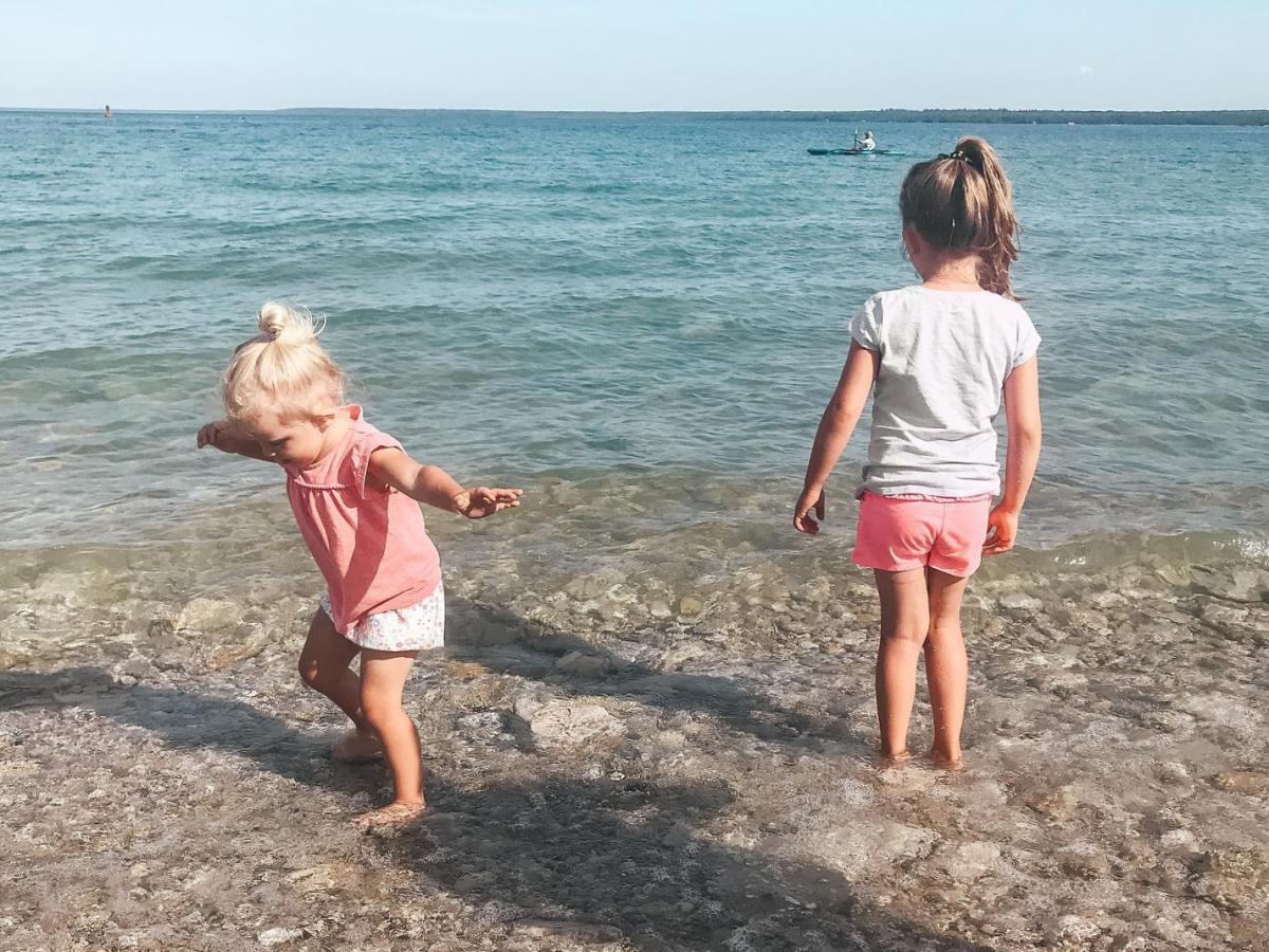 Two young girls wade into the water off Mackinac Island and watch a kayaker in the distance