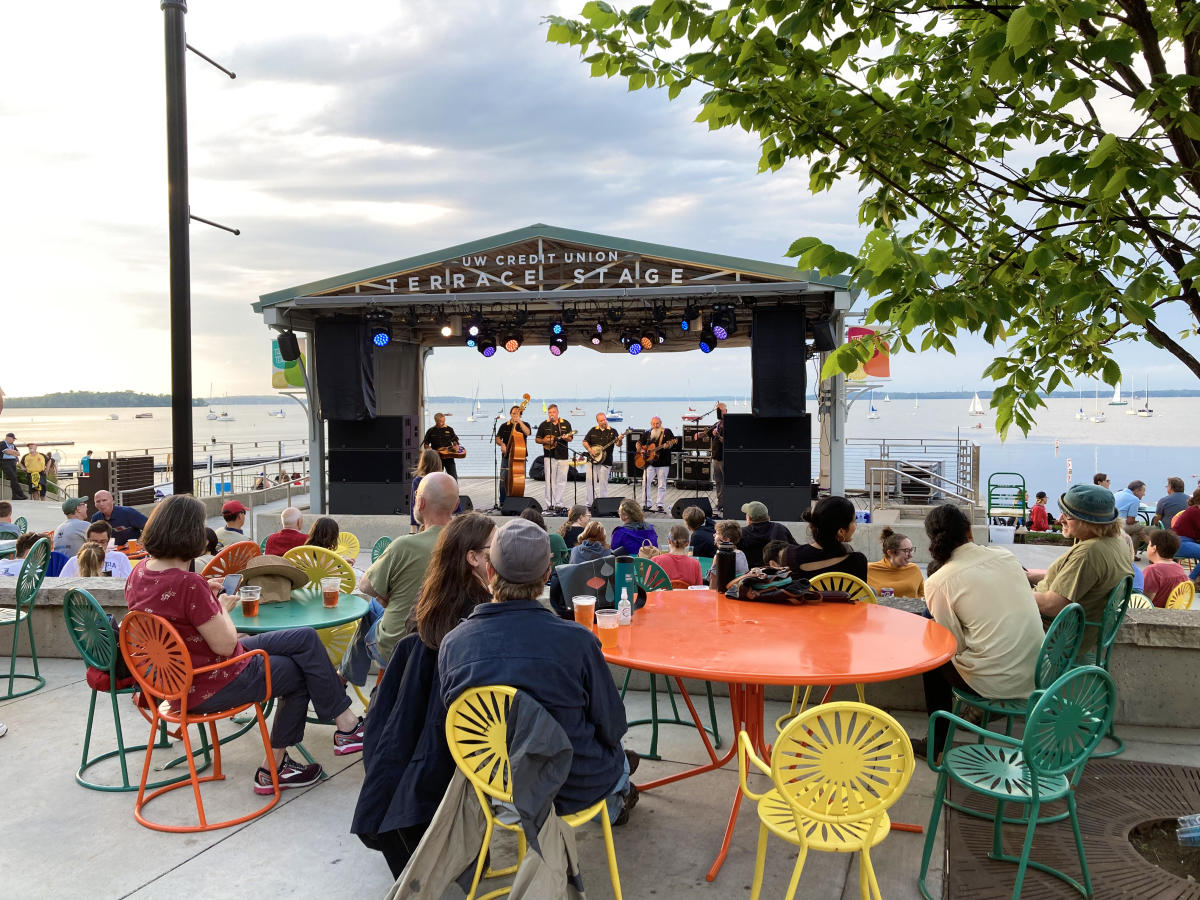 Dozens of people watching a concert at Memorial Union Terrace, sitting on the popular orange, yellow, and green terrace chairs. The people are facing the stage which is in front of Lake Mendota in the background on a sunny Summer day.