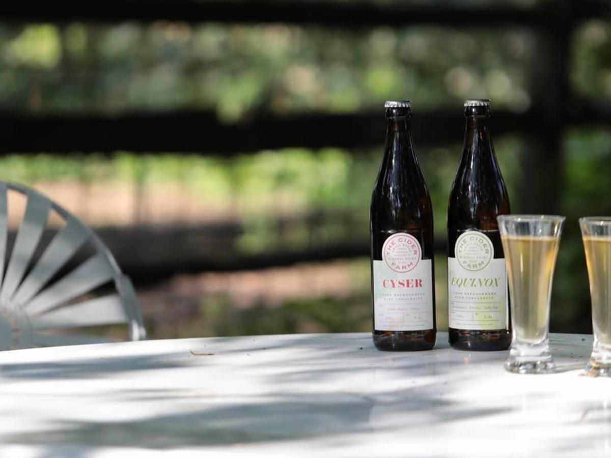 A bottle of Cider Farm Equinox and Cider Farm Cyser sitting with two filled glasses on a white table outside.