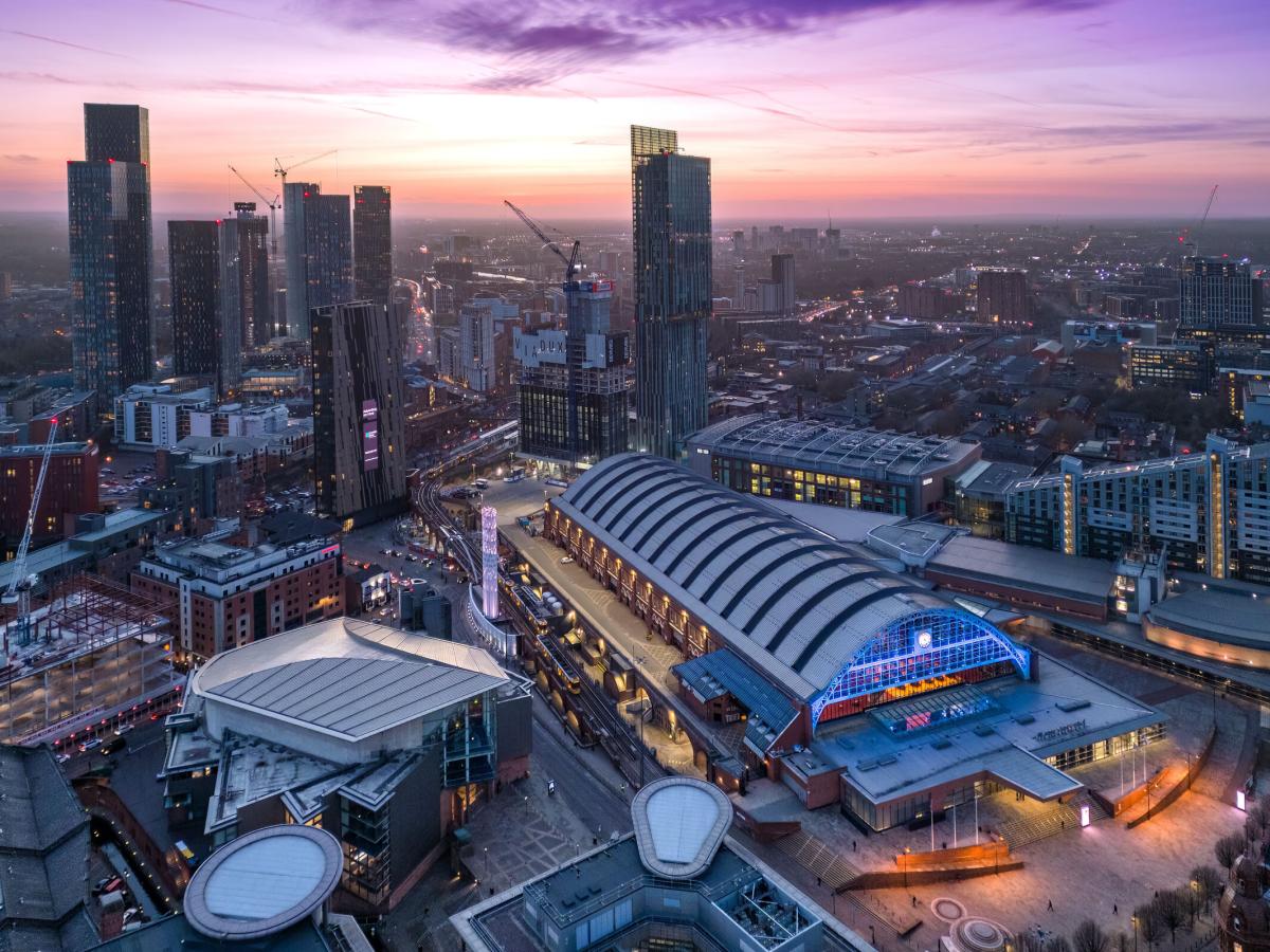 Aerial view of Manchester city centre at sunset, featuring Manchester Central with its arched roof in the foreground and modern skyscrapers rising behind against a pink and purple sky.