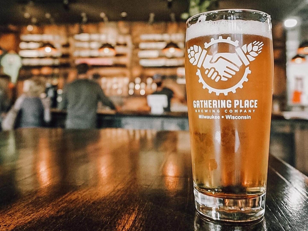 A pint glass of amber beer from Gathering Place Brewing Company sits on a polished wooden table, with a warmly lit taproom and blurred patrons in the background.