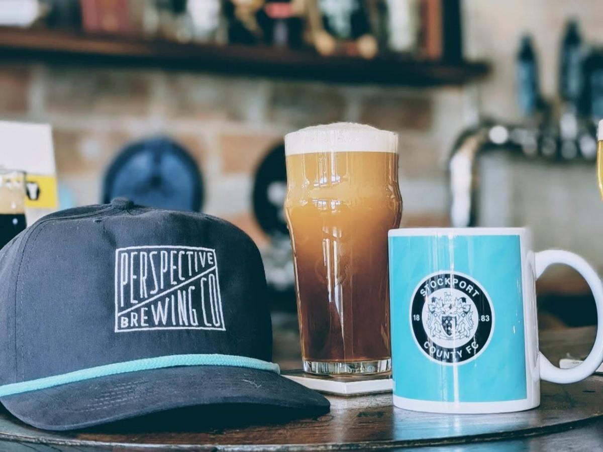 A pint of dark, frothy beer from Perspective Brewing Co. sits on a wooden surface alongside a branded baseball cap and a white and blue coffee mug, with a bar setting and beer taps in the background.