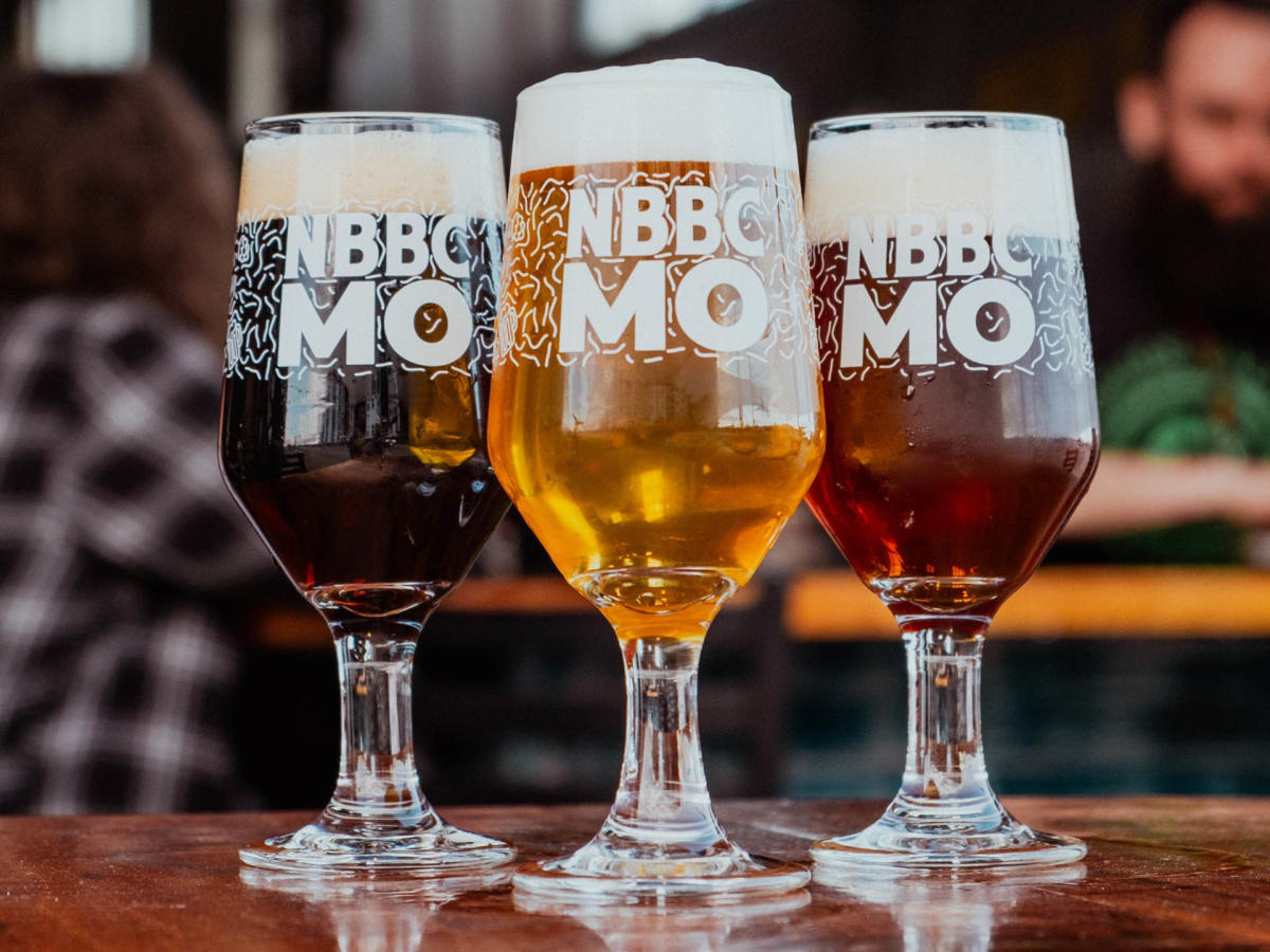Three full beer glasses, filled to the brim with foam at the top of each one, on a wooden table tap and people eating and drinking in the background.