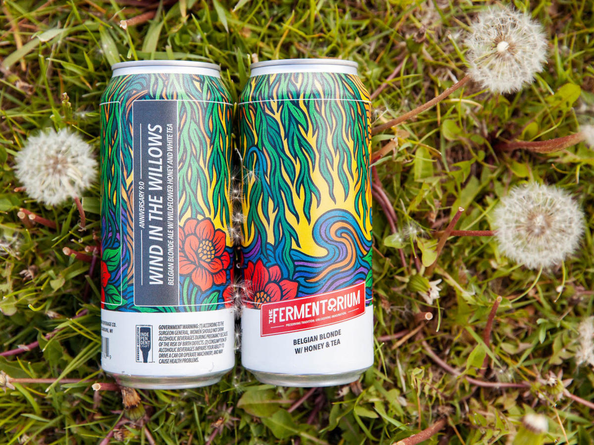 Two colorful cans of “Wind in the Willows” beer from Fermentorium Brewery rest on grass among blooming dandelions, featuring vibrant artwork with flames, flowers, and swirling patterns.