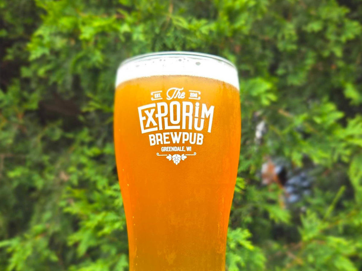A pint glass filled with golden-orange beer from The Explorium Brewpub, set against a backdrop of green foliage.