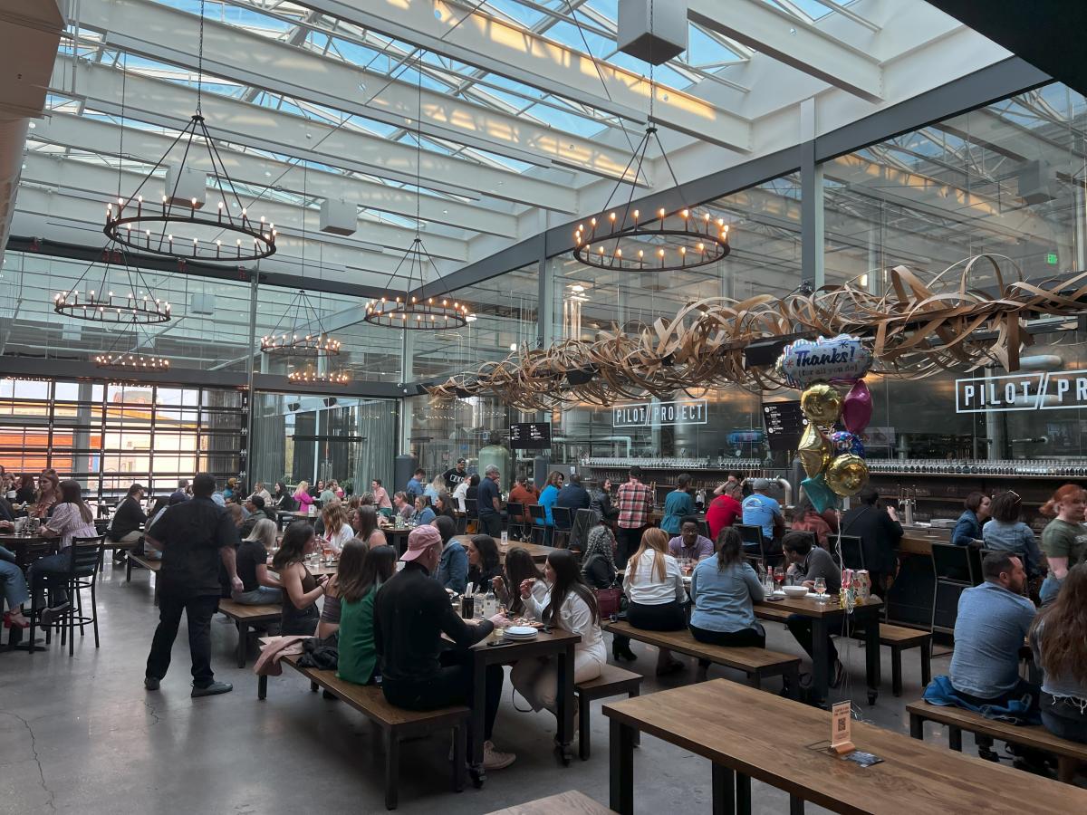 A large indoor beer hall filled with people seated at long wooden tables and bar seating. The space has a high glass ceiling with round chandeliers and a wall of windows letting in natural light. The Pilot Project bar area runs along the right side with taps visible in the background. Decorative wooden ribbon art hangs above the bar. A cluster of thank you balloons sits on one table while groups of guests eat, talk and enjoy drinks throughout the busy space.