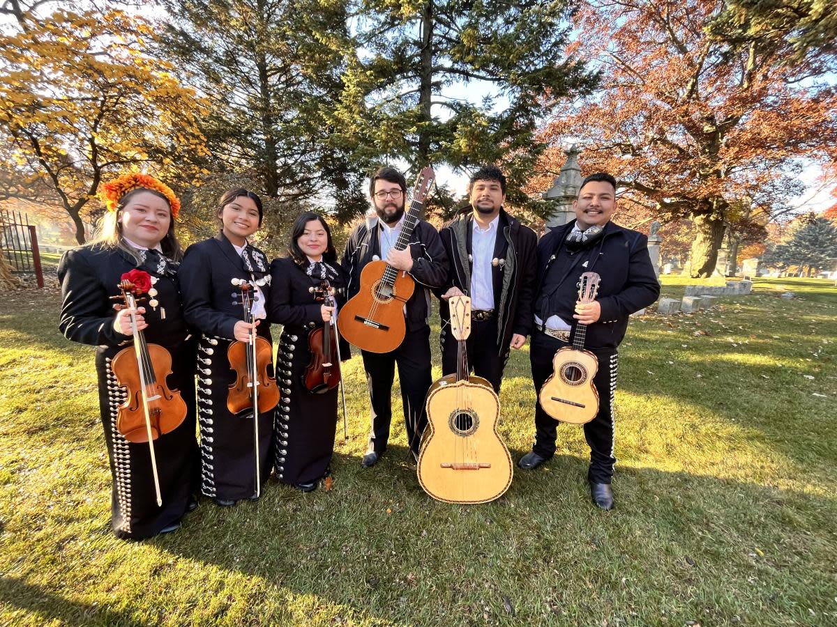 A mariachi group of six musicians poses outdoors on a sunny autumn day, holding violins, guitars, and a vihuela. The women wear traditional black mariachi suits with embroidered details, while the men wear black suits with ties or sashes. Fall trees with orange leaves create a colorful backdrop.