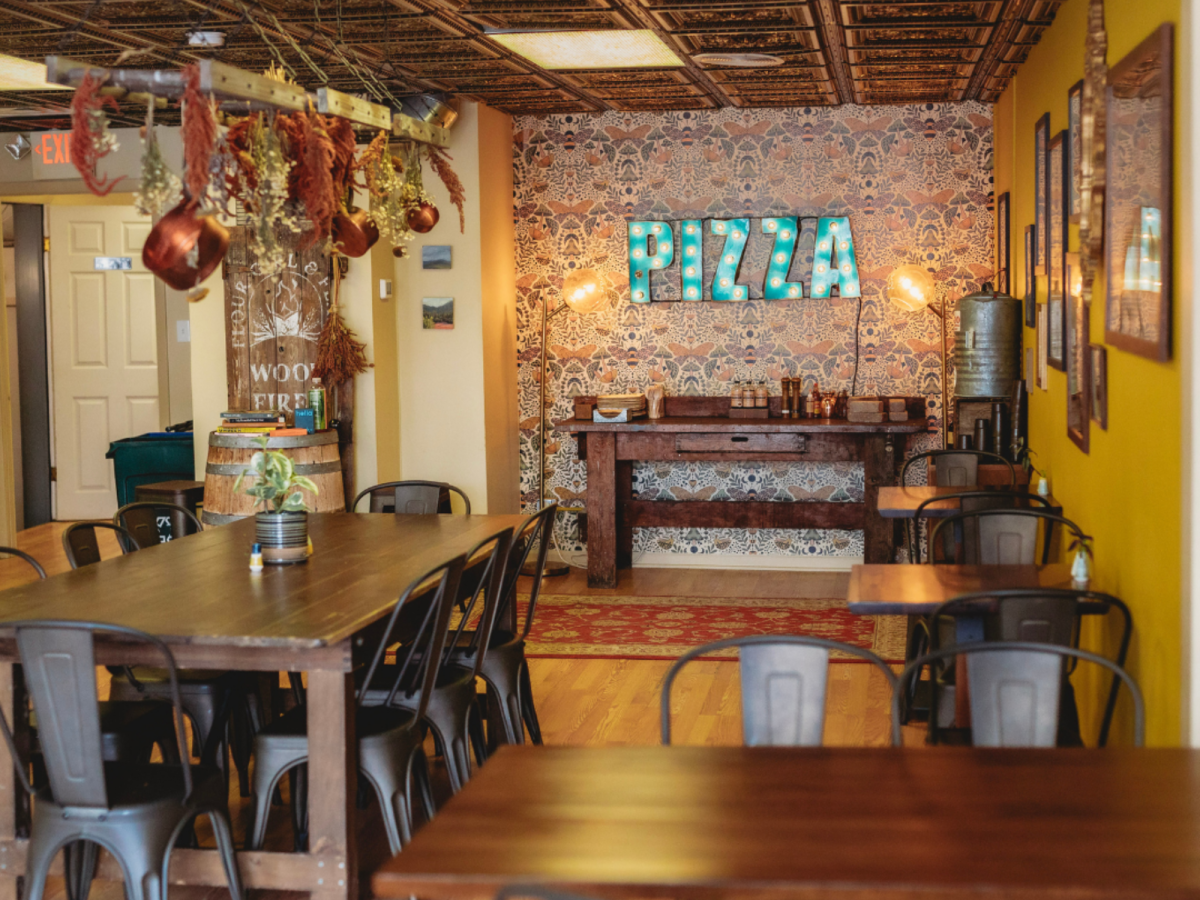 Interior of Flour Girl & Flame with wooden tables, metal chairs, hanging copper pots and a decorative wall with a bright “PIZZA” sign.