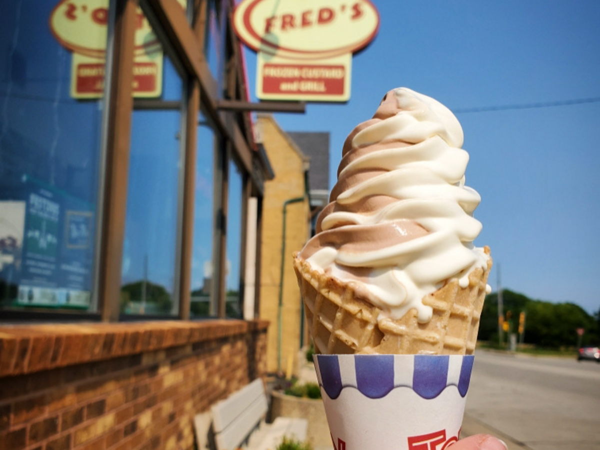 A chocolate-and-vanilla twist frozen custard cone is held outside Fred's Frozen Custard & Grill, with the restaurant's sign and storefront softly blurred in the background on a sunny day.
