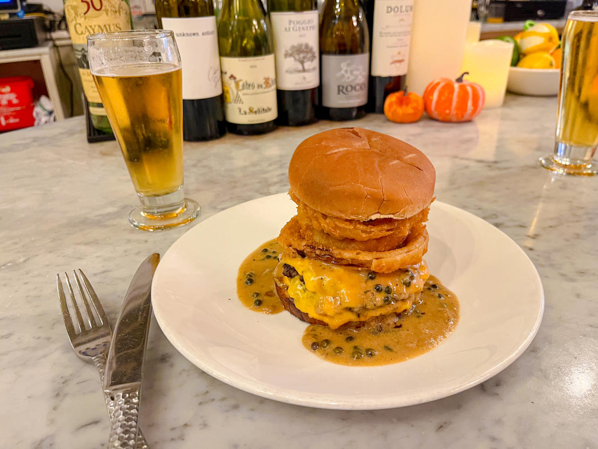A gourmet burger stacked with melted cheese, crispy onion rings, and creamy peppercorn sauce, served on a marble countertop with a glass of beer and bottles of wine in the background.