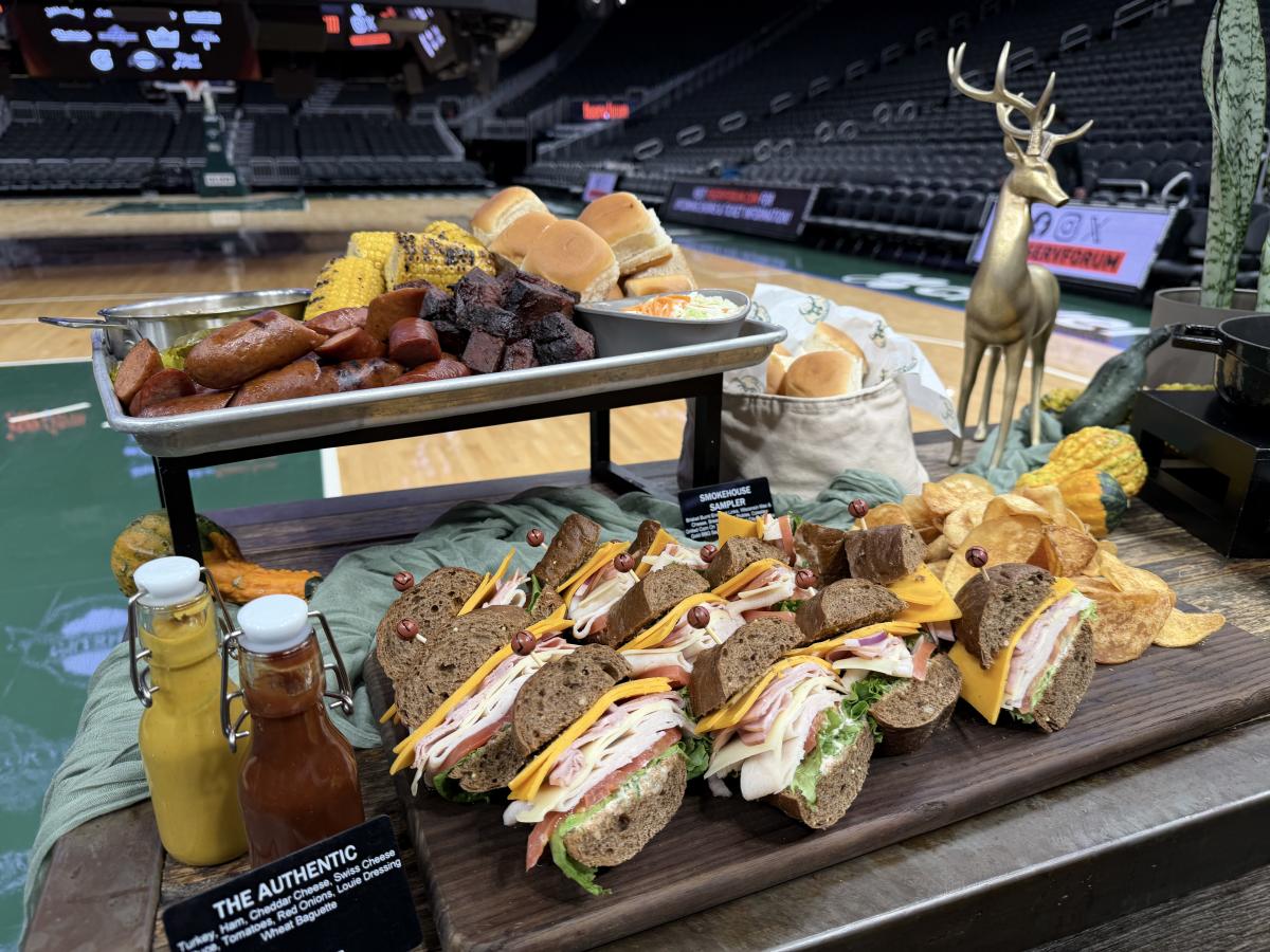 A spread of Fiserv Forum food offerings, including smoked meats, sausages, corn on the cob, deli sandwiches, rolls and chips, displayed courtside at the home of the Milwaukee Bucks.
