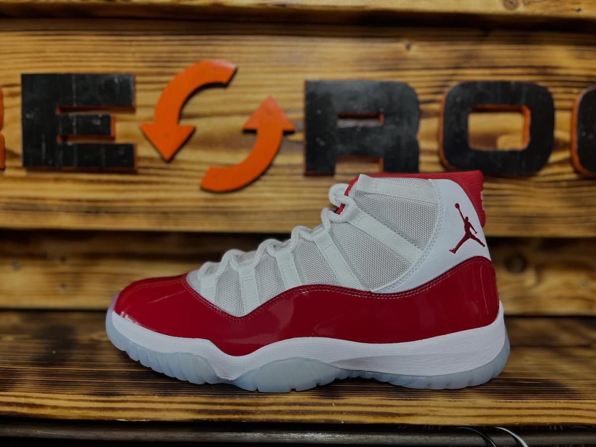 A red-and-white Air Jordan sneaker is displayed on a wooden counter inside Rerocc, with the store's logo mounted on a wood-paneled wall in the background. The high-top shoe is shown in profile, highlighting its red patent leather accents and white mesh upper.