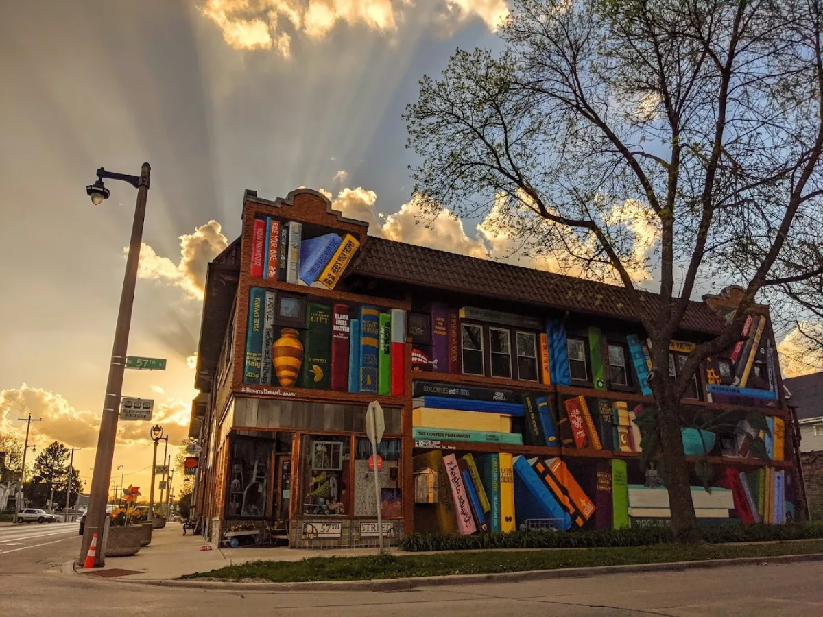Corner bookstore building covered in a colorful mural of oversized books, photographed at sunset with golden light streaming through clouds behind it. The two story brick structure sits at an intersection, with a street sign visible and a sidewalk wrapping around the corner. Painted book spines of various colors and titles line the exterior walls, creating the look of a stacked library across the façade, while a leafless tree frames the right side of the scene.
