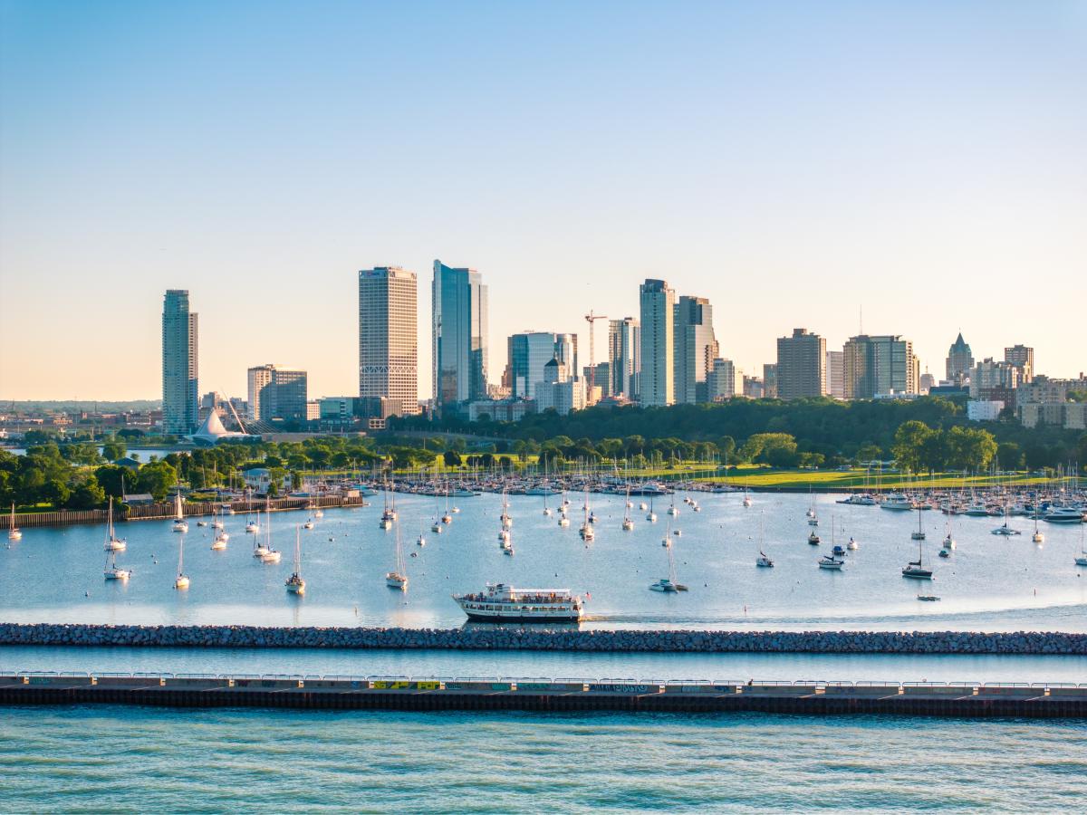 A wide view of Milwaukee’s lakefront skyline on a clear day, with dozens of sailboats anchored in a calm marina and a tour boat cruising along the breakwater in the foreground. Green parkland lines the waterfront beneath a cluster of downtown high-rise buildings and glass towers.