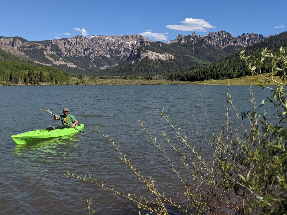 Man paddling a bright green tandem kayak across Silver Jack Reservoir, a high elevation body of water with the jagged Cimarron Mountains on the horizon, on a sunny day in the summertime.