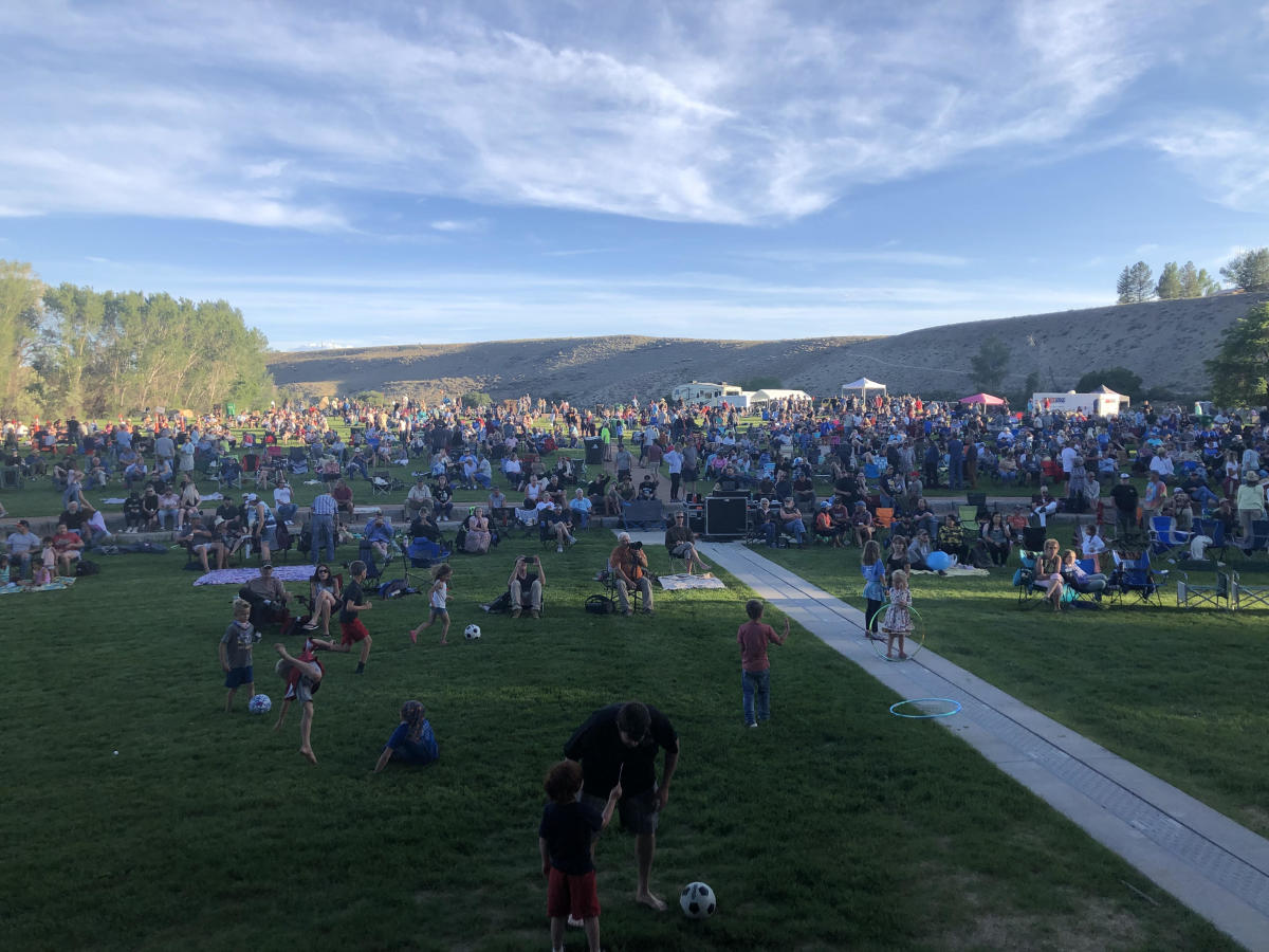 A crowd of children playing, dogs, and adults in lawn chairs and on blankets stare ahead at the Montrose Rotary Amphitheater during the Montrose Summer Music Series 2022